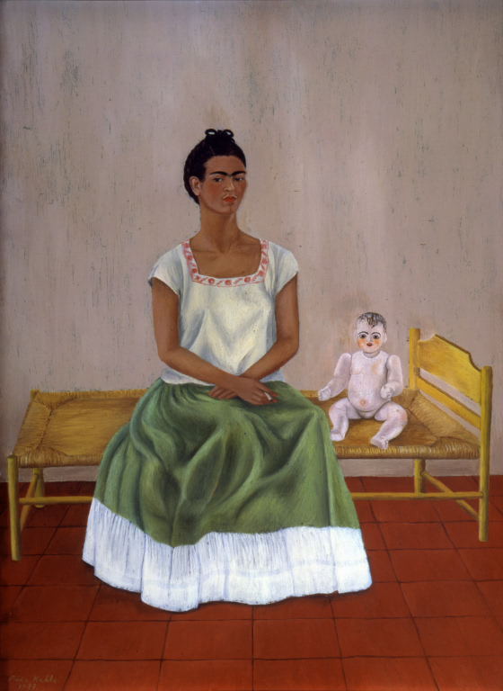 Kahlo sits on a small bed next to a pale, stiff baby doll. Wearing a white top and green skirt, she stares directly out of the frame at the viewer.
