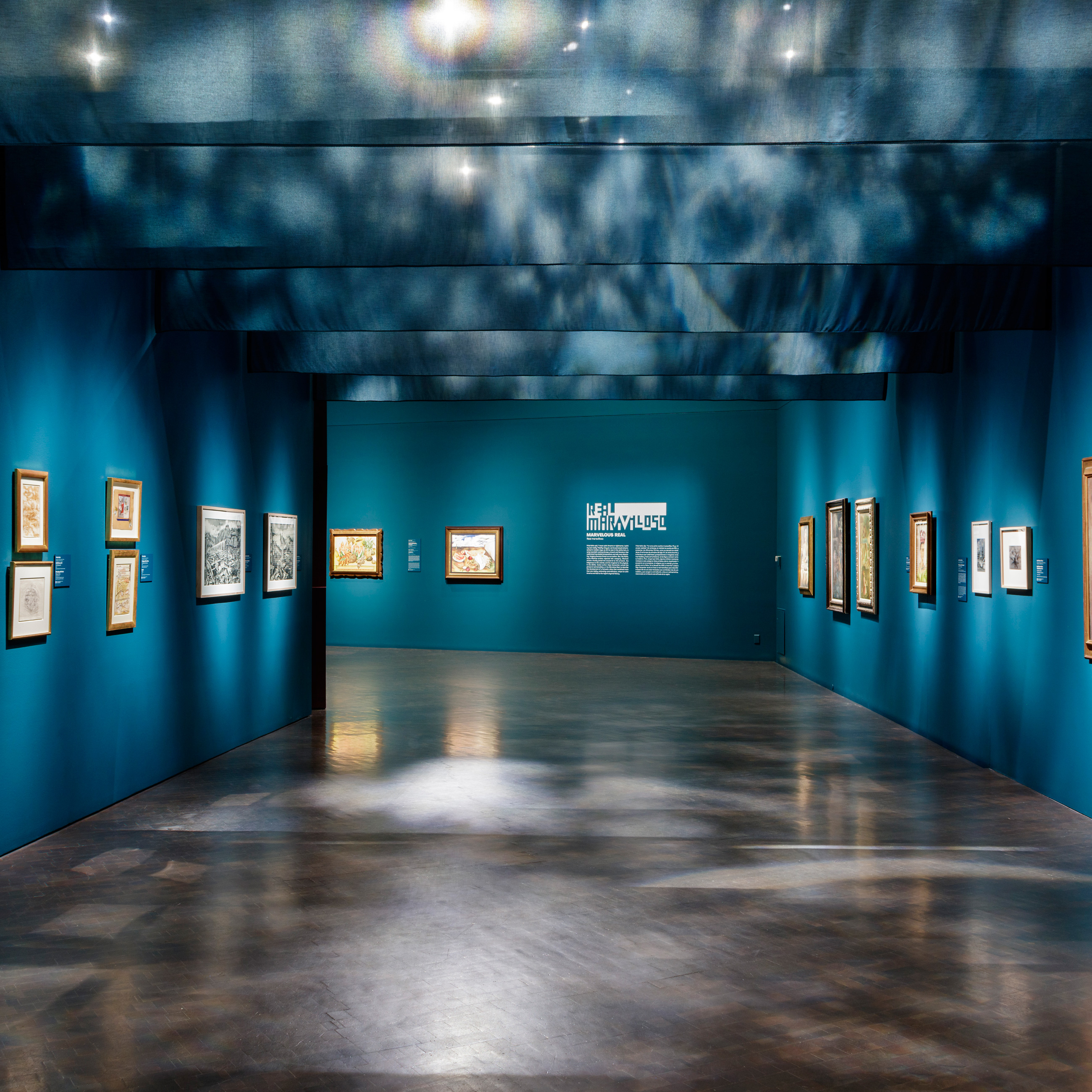 photo of one of the galleries in the Mexican Modernism exhibition featuring paintings on dark teal walls and gauzy cloth hanging from the ceiling casting light and shadows
