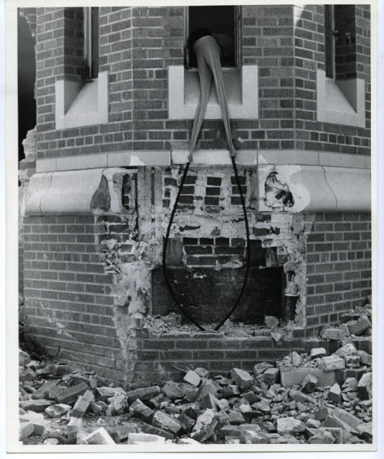 Black and white photograph of Nengudi leaning out the window of a crumbling building. She wears pantyhose on her head attached to two long pieces of wire.