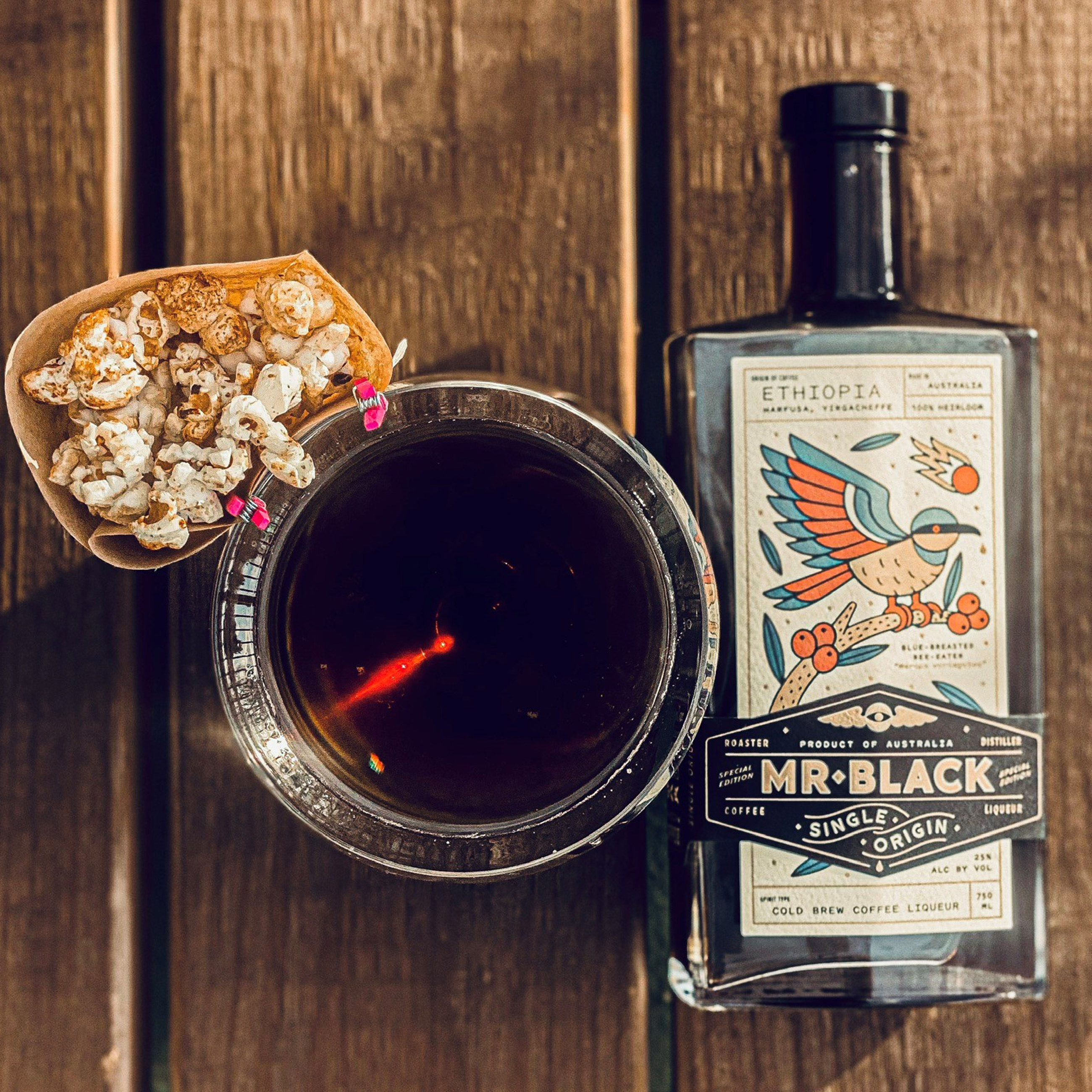 picture of popcorn, Black as Knight cocktail, and a bottle of Mr. Black coffee liquer lying on a wood table