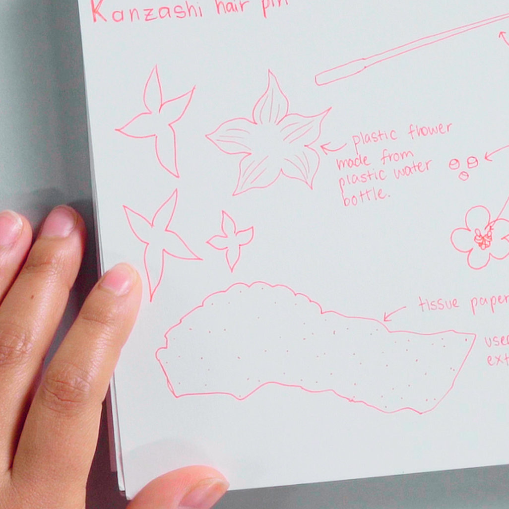 A pair of hands draws jewelry design concepts in pink pen on a sketchbook.