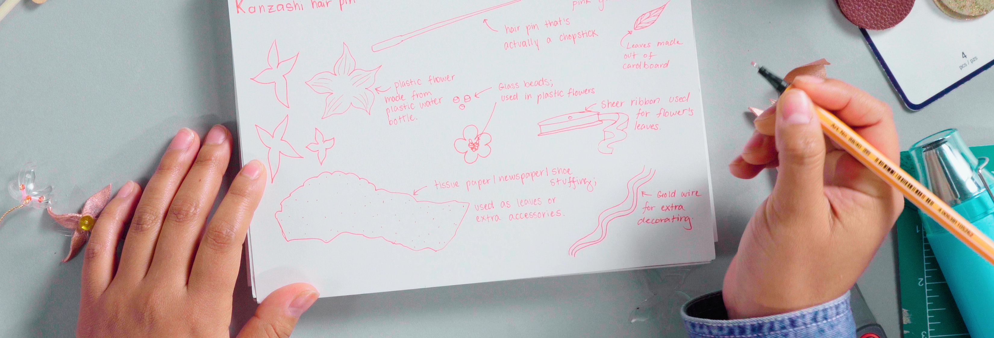 A pair of hands draws jewelry design concepts in pink pen on a sketchbook.