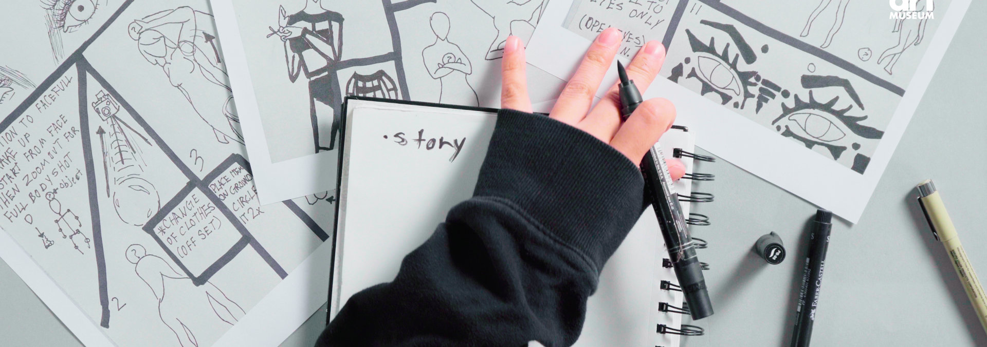 A pair of hands holding a brush pen and a sketchbook on a grey table covered in story-book style drawings.