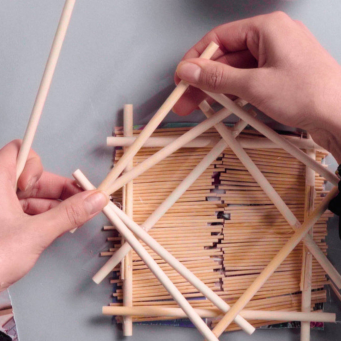 Photo of a pair of hands crafting a multi-level structure out of wodden dowels and other craft materials on a grey desk covered in similar craft materials.