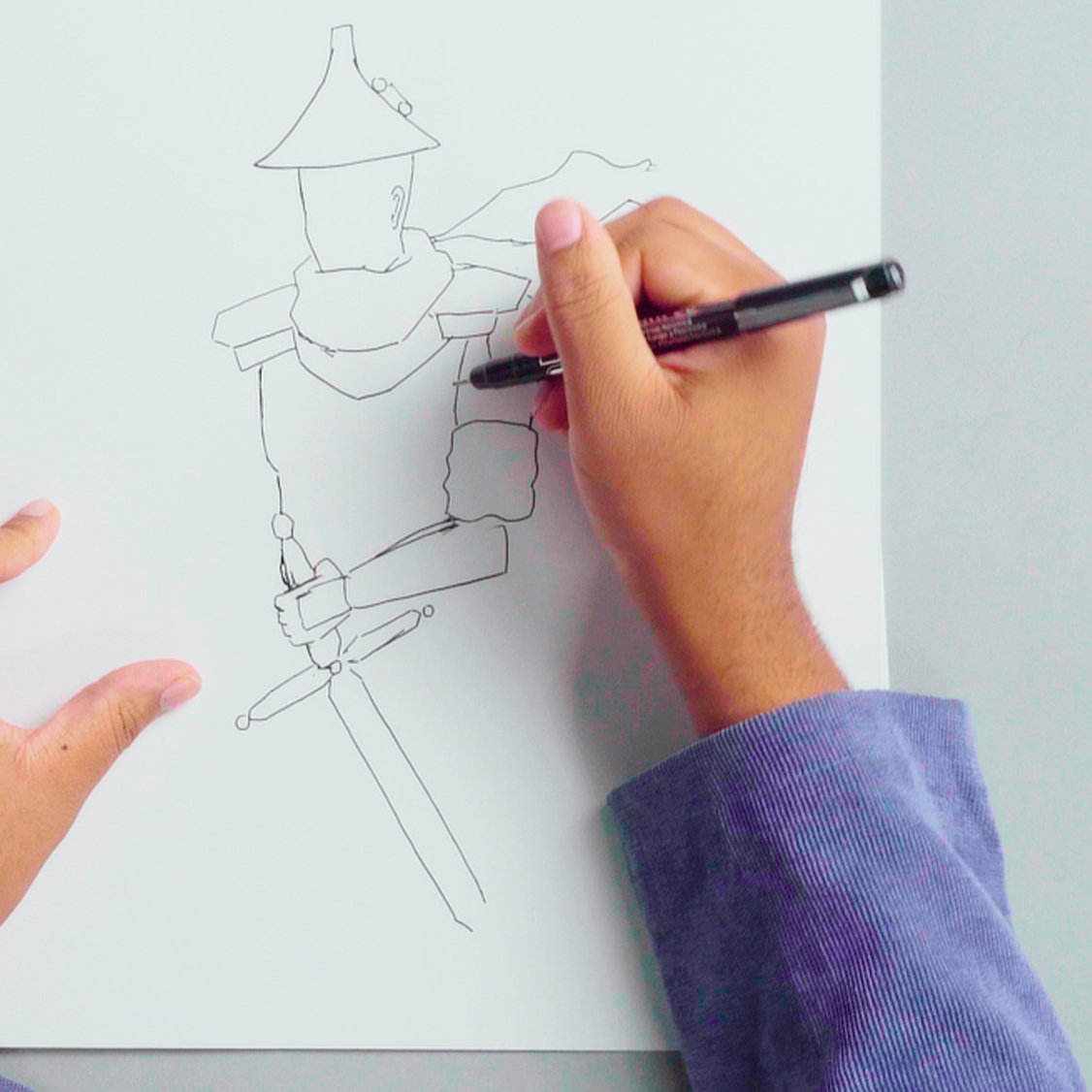 A pair of hands over a table covered in drawing supplies draws a character on a clean sheet of paper