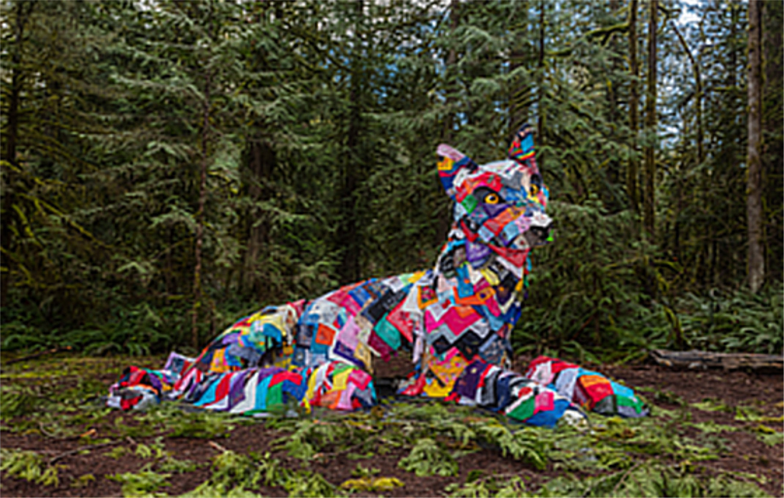 Giant canine sculpture covered in a colorful patchwork of bandanas, set amongst pine trees.