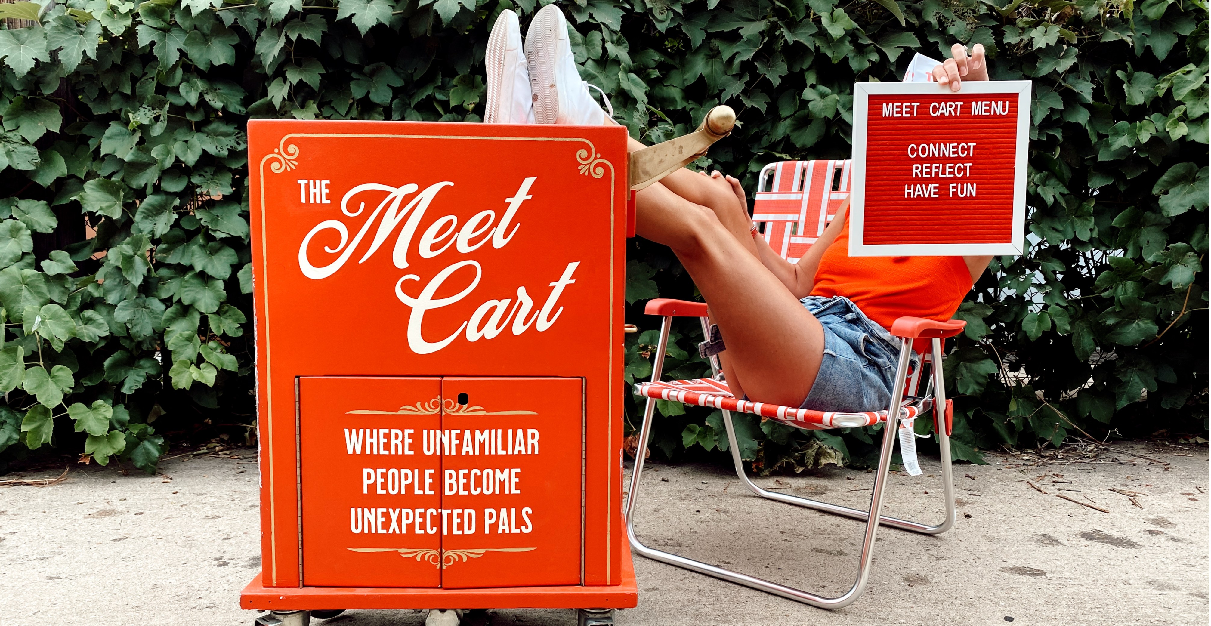 A person sitting on a red lawn chair with feet propped up on The Meet Cart. The cart reads, "The Meet Cart: Where unfamiliar people become unexpected pals." The person is also holding a sign that says, "Meet Cart Menu: Connect, Reflect, Have fun."