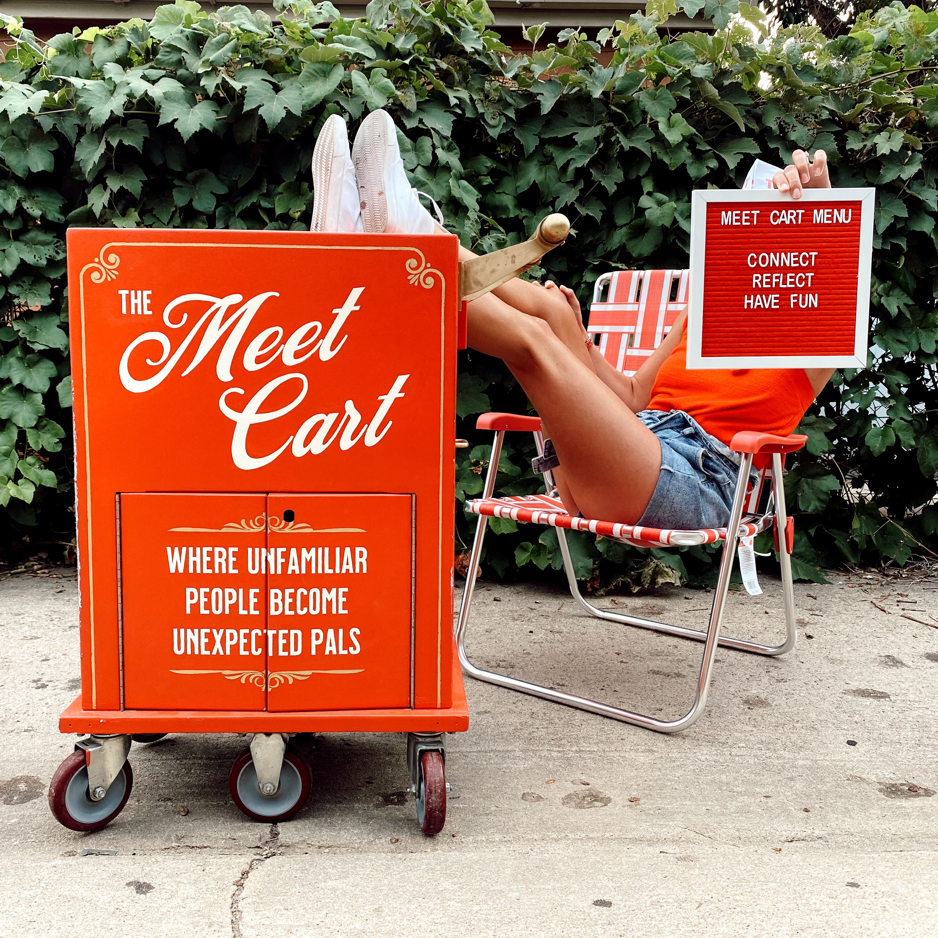 A person sitting on a red lawn chair with feet propped up on The Meet Cart. The cart reads, "The Meet Cart: Where unfamiliar people become unexpected pals." The person is also holding a sign that says, "Meet Cart Menu: Connect, Reflect, Have fun."