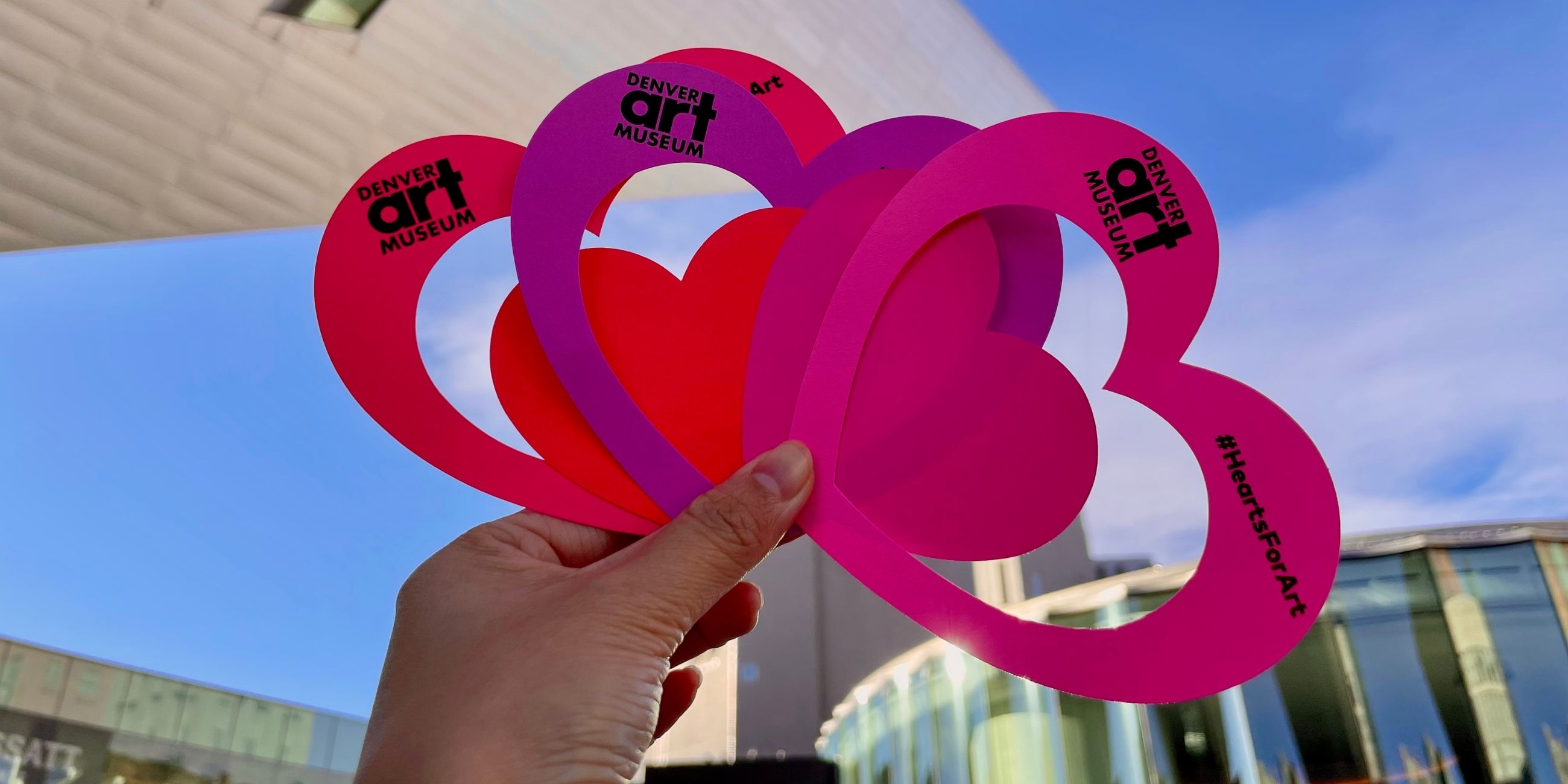 A hand holding paper cut out hearts in front of the Denver Art Museum buildings.