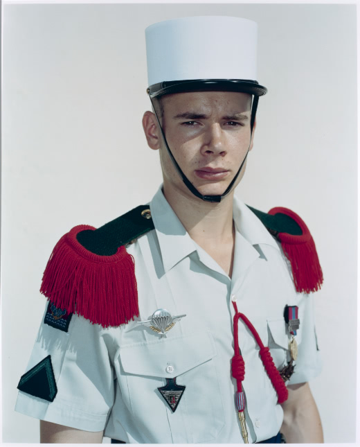 Color portrait of a young man dressed in military parade uniform with a white kepi hat and red epaulettes on his shoulders. He stands against a blank background and has a neutral expression as he looks directly at the camera.