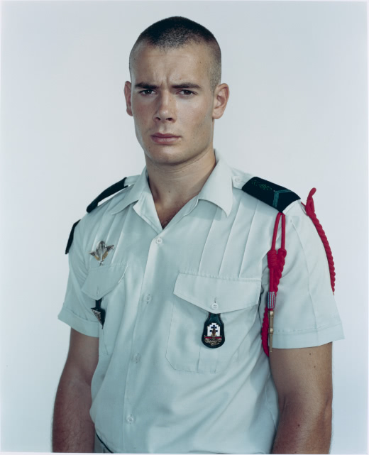 Color portrait of a young male soldier with a buzz cut standing against a blank background. He wears a short-sleeved white uniform and has a neutral expression as he looks directly at the camera.