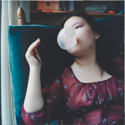 A color portrait of a young woman who leans back on a blue-cushioned chair and blows a bubble as she gazes to the left of the frame. Her right hand holds a strand of her long hair.