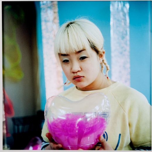A color portrait of a young woman with bleached hair with thick bangs and multiple short braids. She wears a faded yellow sweatshirt and holds a clear plastic heart filled with magenta-pink fuzz.