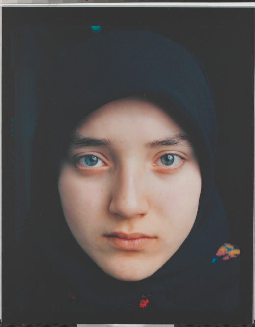 Headshot of a young girl in a black hijab. Small but colorful embroidery is visible under her chin and in the right-hand side of the frame on her shoulder. She looks directly into the camera with blue eyes and the smirk of a smile.