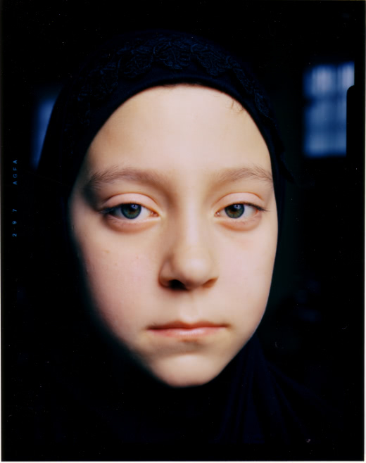 Headshot of a young girl in a black embroidered hijab. She looks directly into the camera with striking greenish-hazel eyes. Her mouth is set in a flat line.