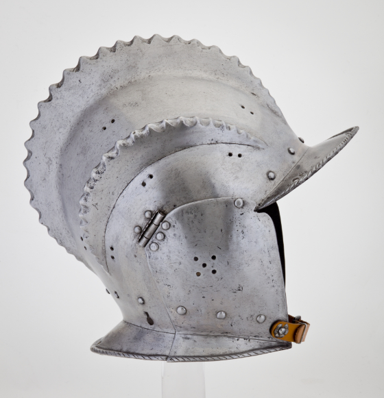 A metal helmet with an open face, short visor, leather chin strap, and three fin-like ridges running from the front to the back.