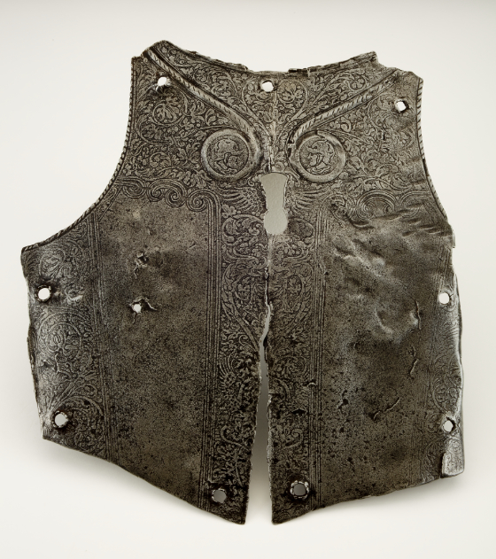 A flattened metal chest plate with decorative engravings through the center and perimeter. The chest plate has a “keyhole” cut out in the center with nine smaller round holes along the perimeter of the plate.