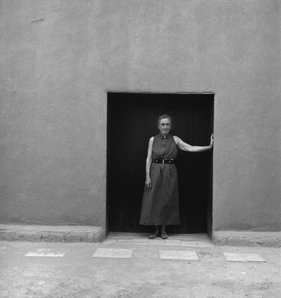 A black and white full-length portrait of O’Keeffe standing in the doorway of an adobe building with one hand resting against the doorway.