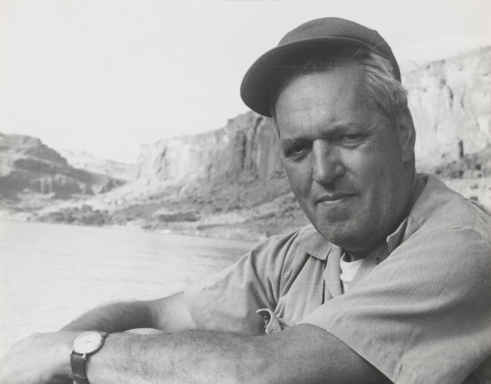 A black and white portrait of Todd Webb from the chest up sitting in front of a river. He looks towards the camera and wears a baseball cap.