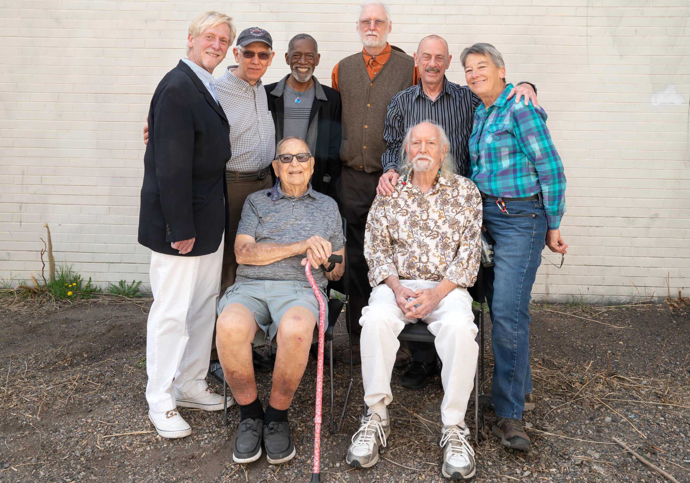 Eight artists are in front of an off-white wall. Two are sitting in chairs in in front of the other six. The six in the back have their arms around each other.