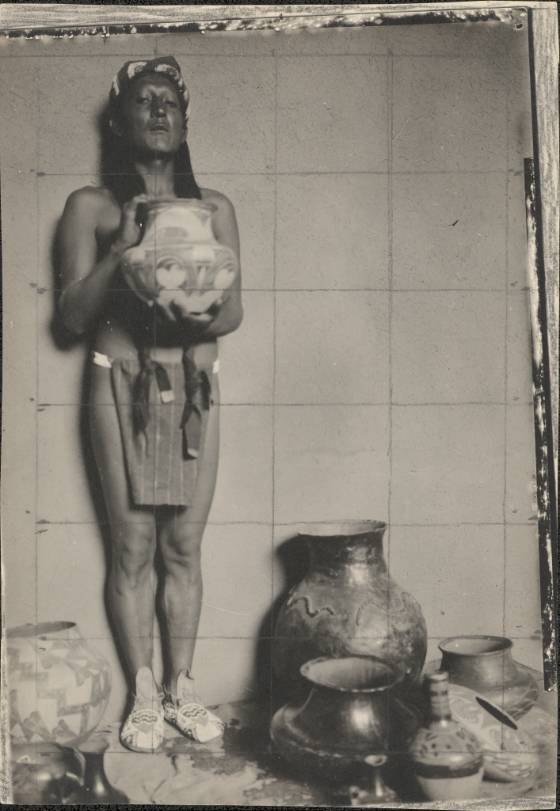 Black and white photo of Jerry Mirabal, an Indigenous man of Taos Pueblo. He poses in a loin cloth and stands rigidly with his back pressed against a wall amid an array of pots. His face is inexpressive and he holds a pot up mid-chest for display.