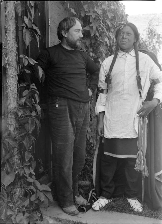 Black and white photo of E. I. Couse, a white artist, and Ben Lujan, an Indigenous man from Taos Pueblo. The two stand side-by-side in front of foliage. Couse, left, looks right toward Lujan, putting his face in profile. Lujan, right, stares straight ahead.