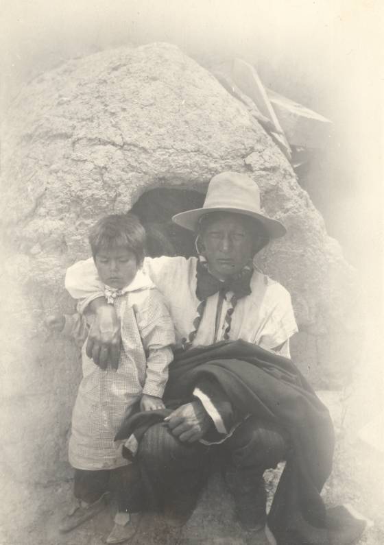 Black and white photo of Jerry Mirabal, right, an Indigenous man from Taos Pueblo, posing with his arm around a young child, left.