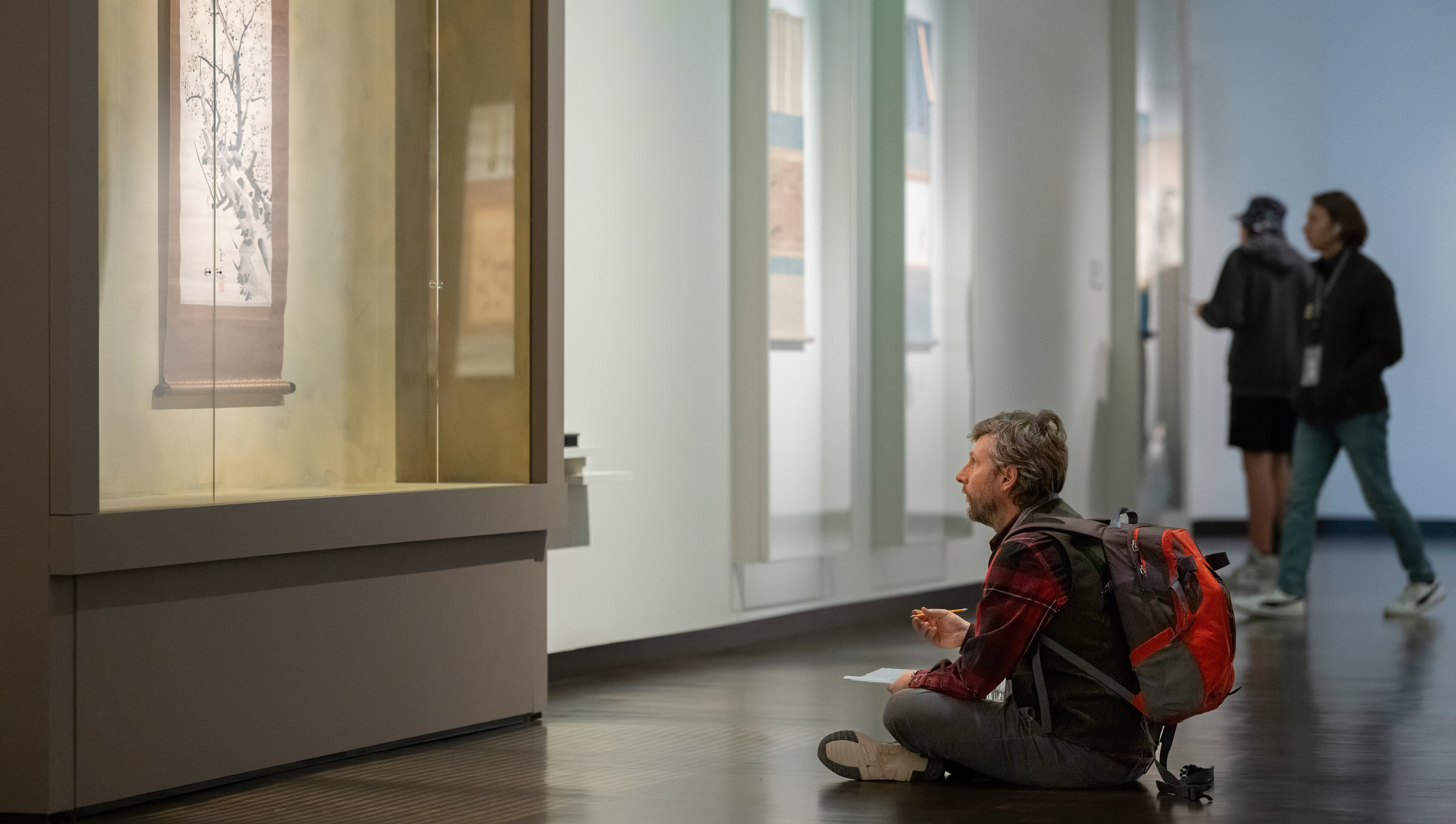 Man sitting down in the galleries looking up to an artwork with a sketchpad