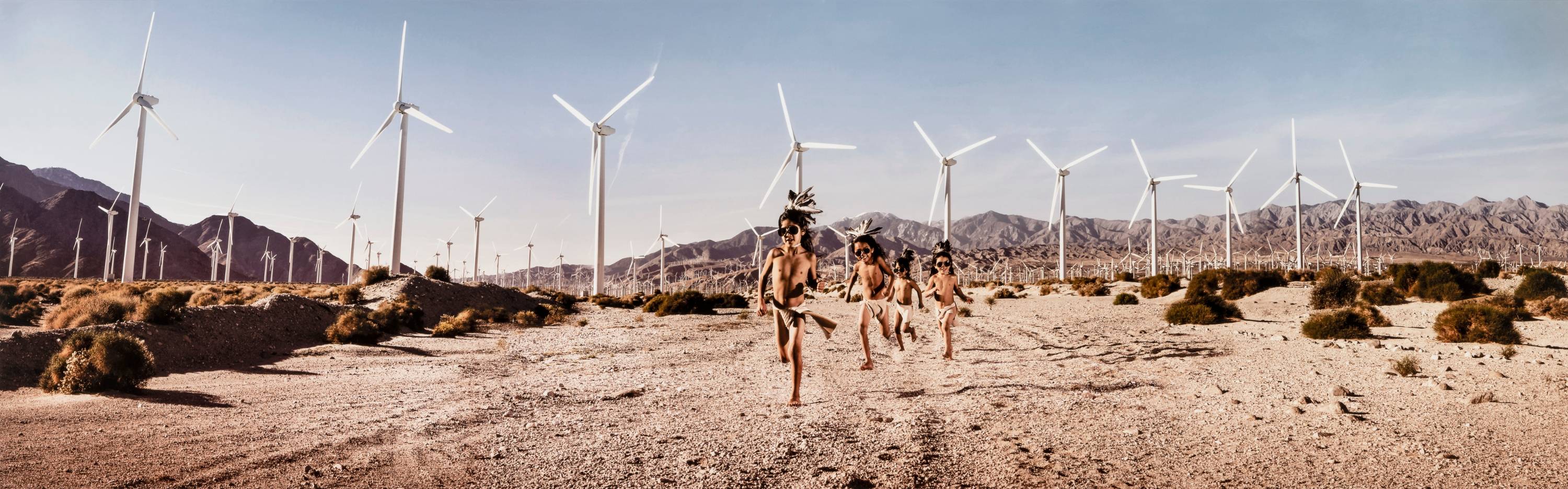 In a desert landscape, four children wear feathers in their hair and loincloths. They run towards the foreground. In the background, wind turbines spin before hazy mountains.