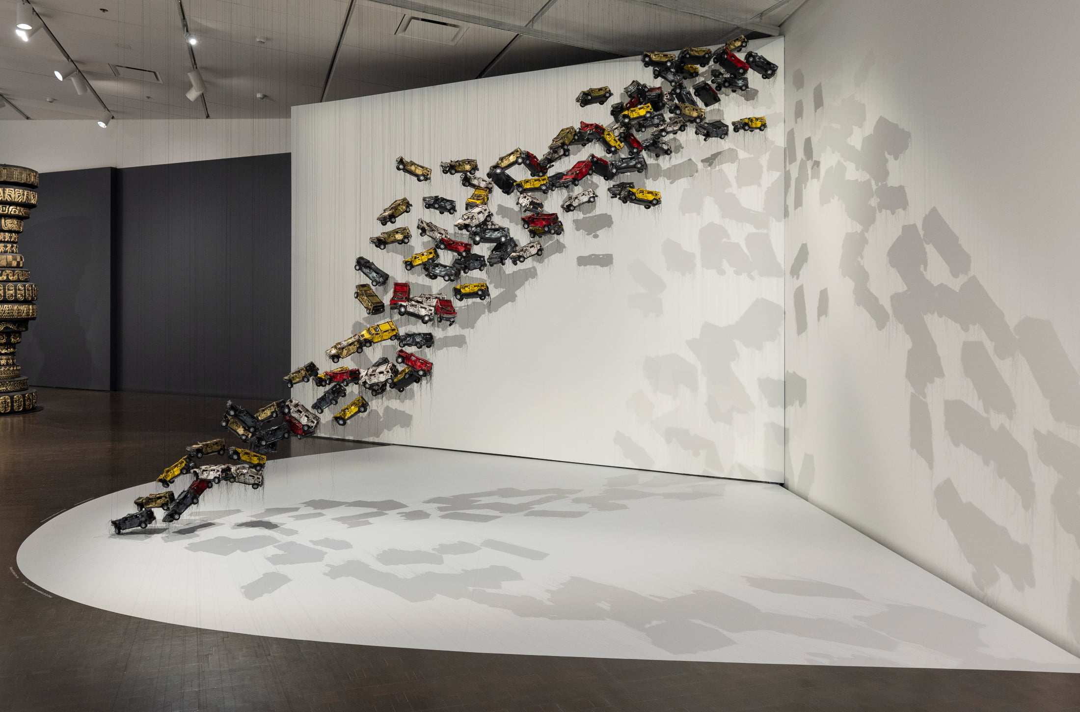 Small, brick-sized Hummers suspended in the air cascade in a diagonal resembling the United States/Mexico border. It goes from ten o’clock on the left, to four o’clock on the right, never touching the floor. The Hummers are different shades of yellow, red, white, grey, and beige.