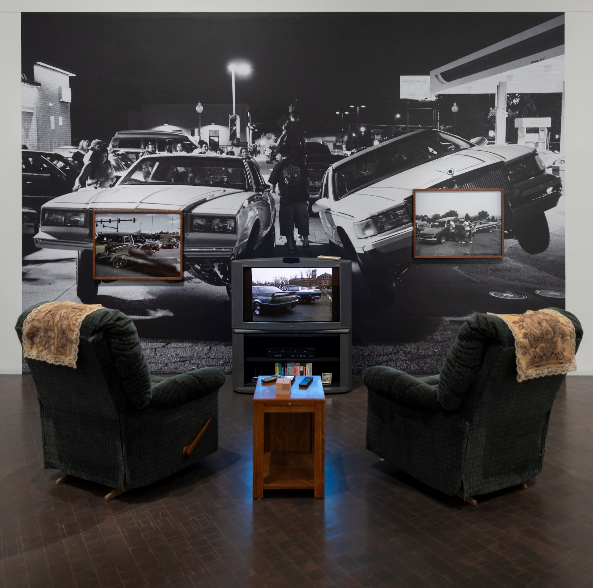Two armchairs, with an end table between them, face a TV which plays videos of lowrider meetups. The wall behind the TV has photos of lowriders from meetups and a wall-sized photo mural ot two lowriders at a meetup.
