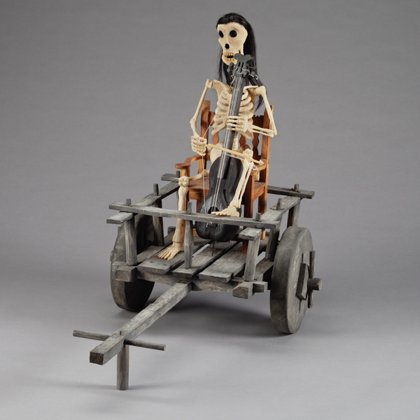 Sculpture of a skeletal man holding an instrument while riding in a wooden cart