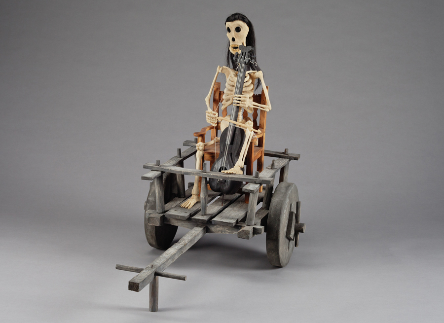 Sculpture of a skeletal man holding an instrument while riding in a wooden cart