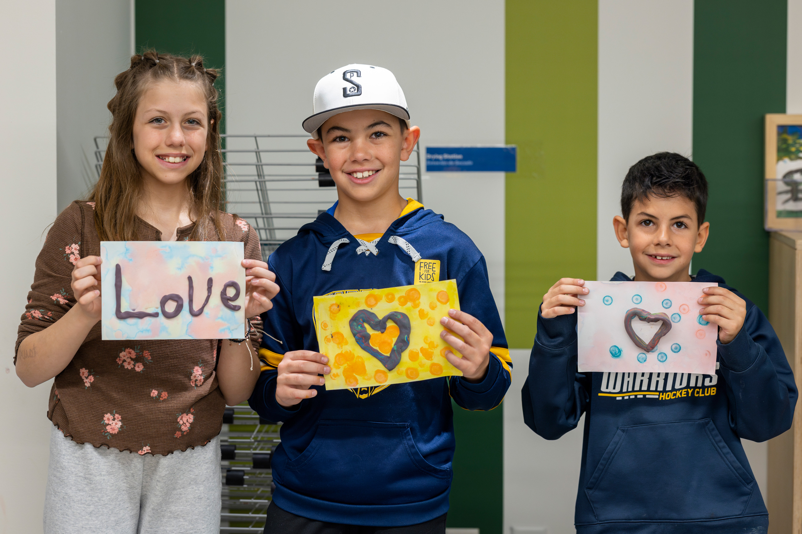 three children hold up artwork, one spells the word love, two are images of hearts