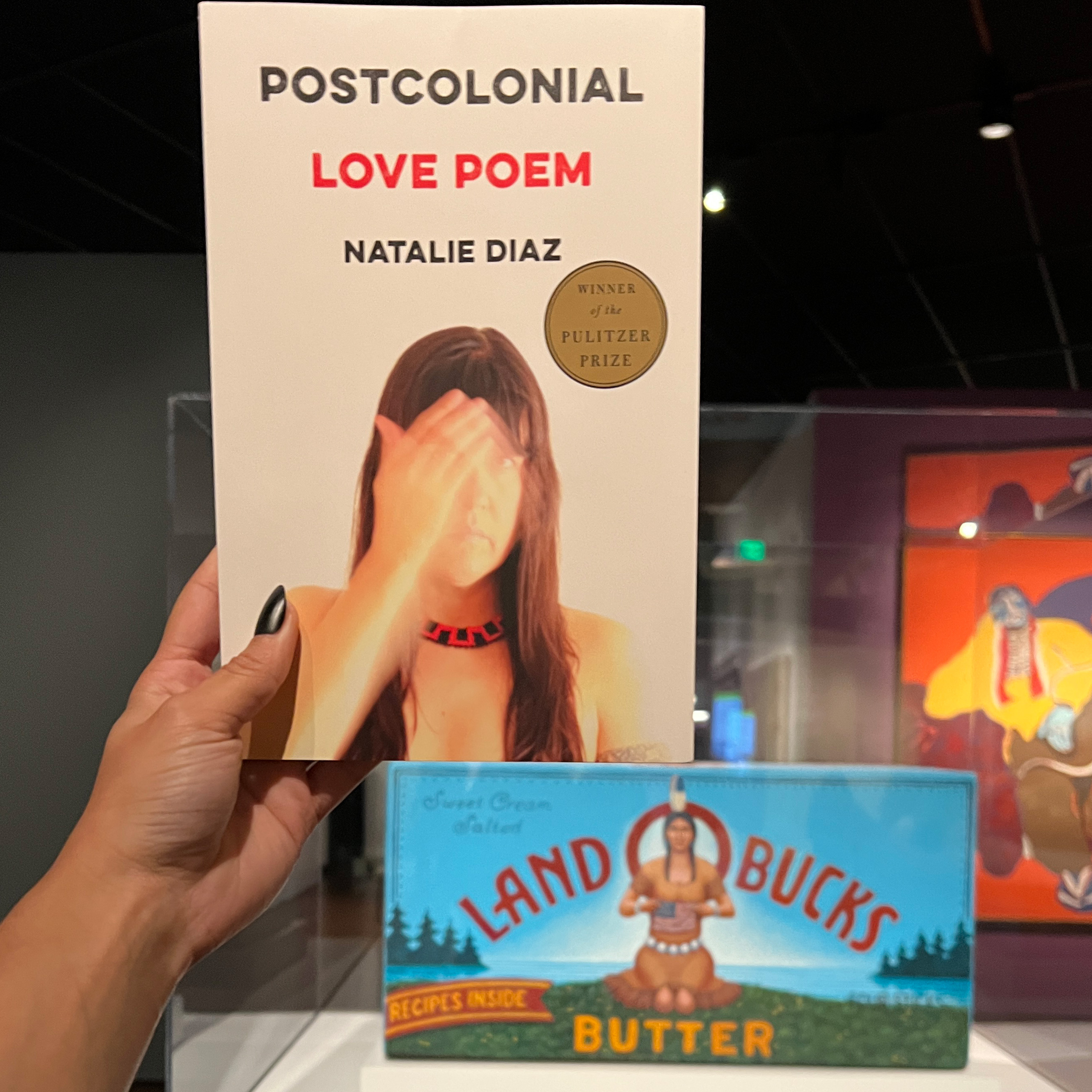 hand holding Postcolonial Love Poems book next to Land o Bucks artwork parodying land of lakes butter in Indigenous gallery