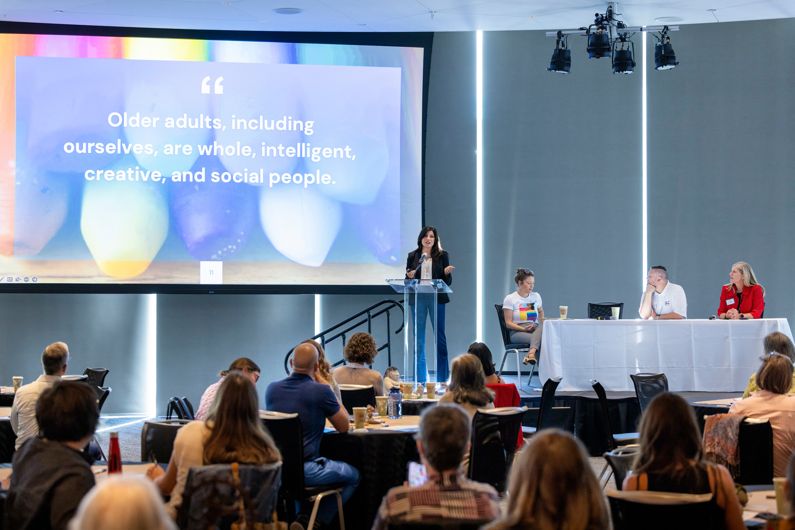 White woman of middle age with brown hair standing and speaking at a podium in front of a large projected image of an older woman and the words, &quot;Creative Aging.&quot;
