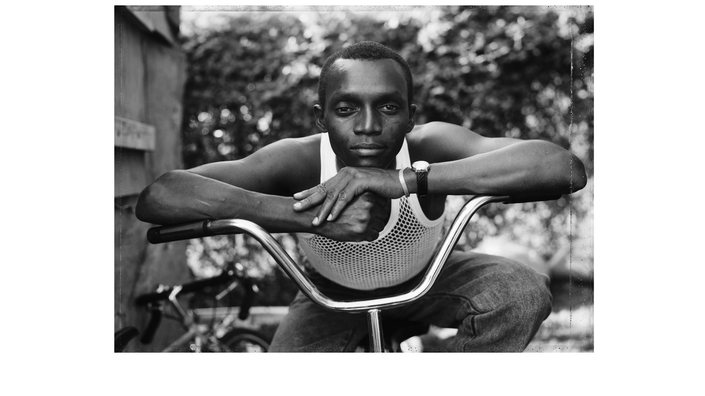 Black and white photograph of a young black man resting on the handles of his bike