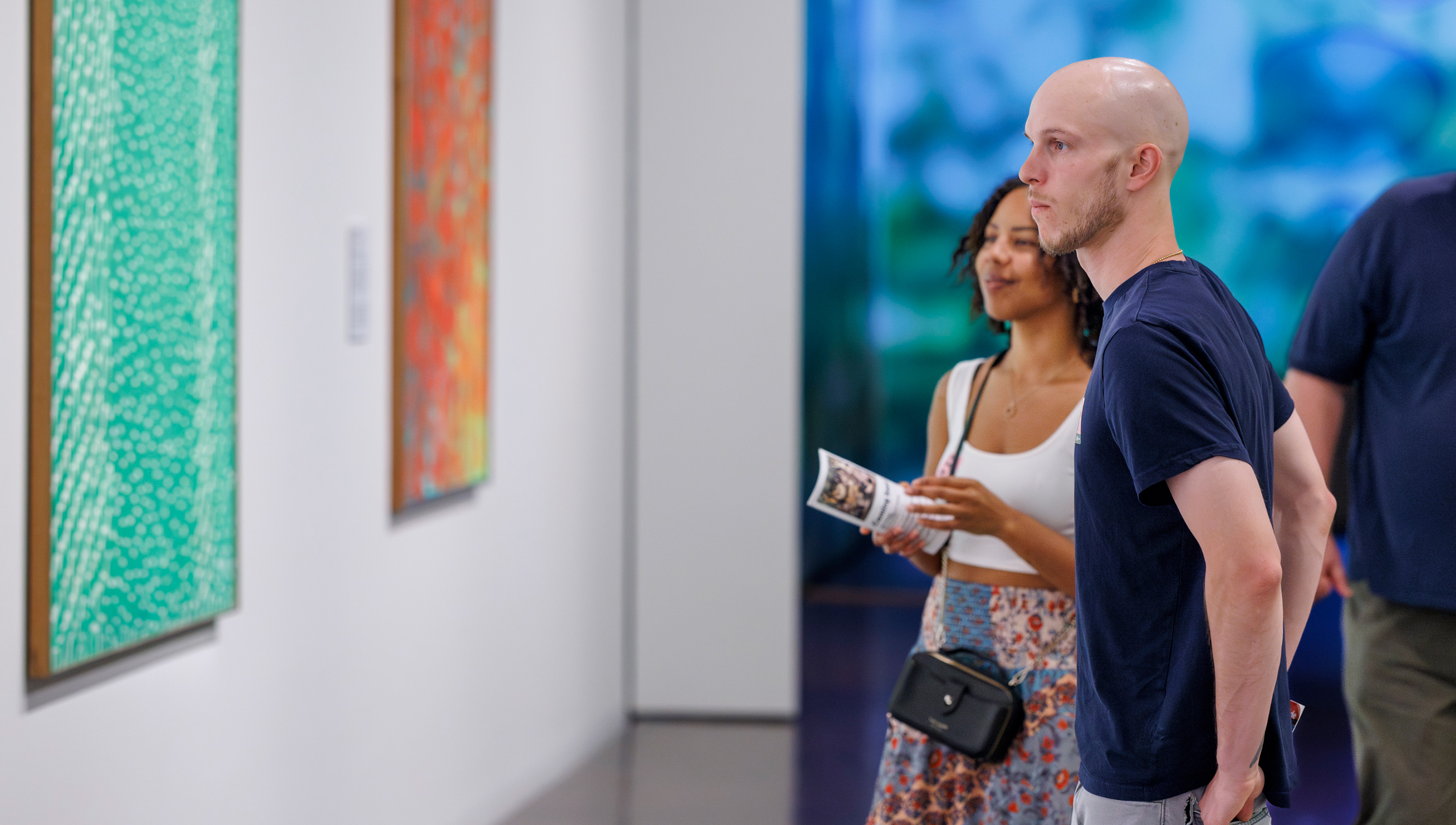 a black woman and a white man look at colorful abstract paintings on a gallery wall