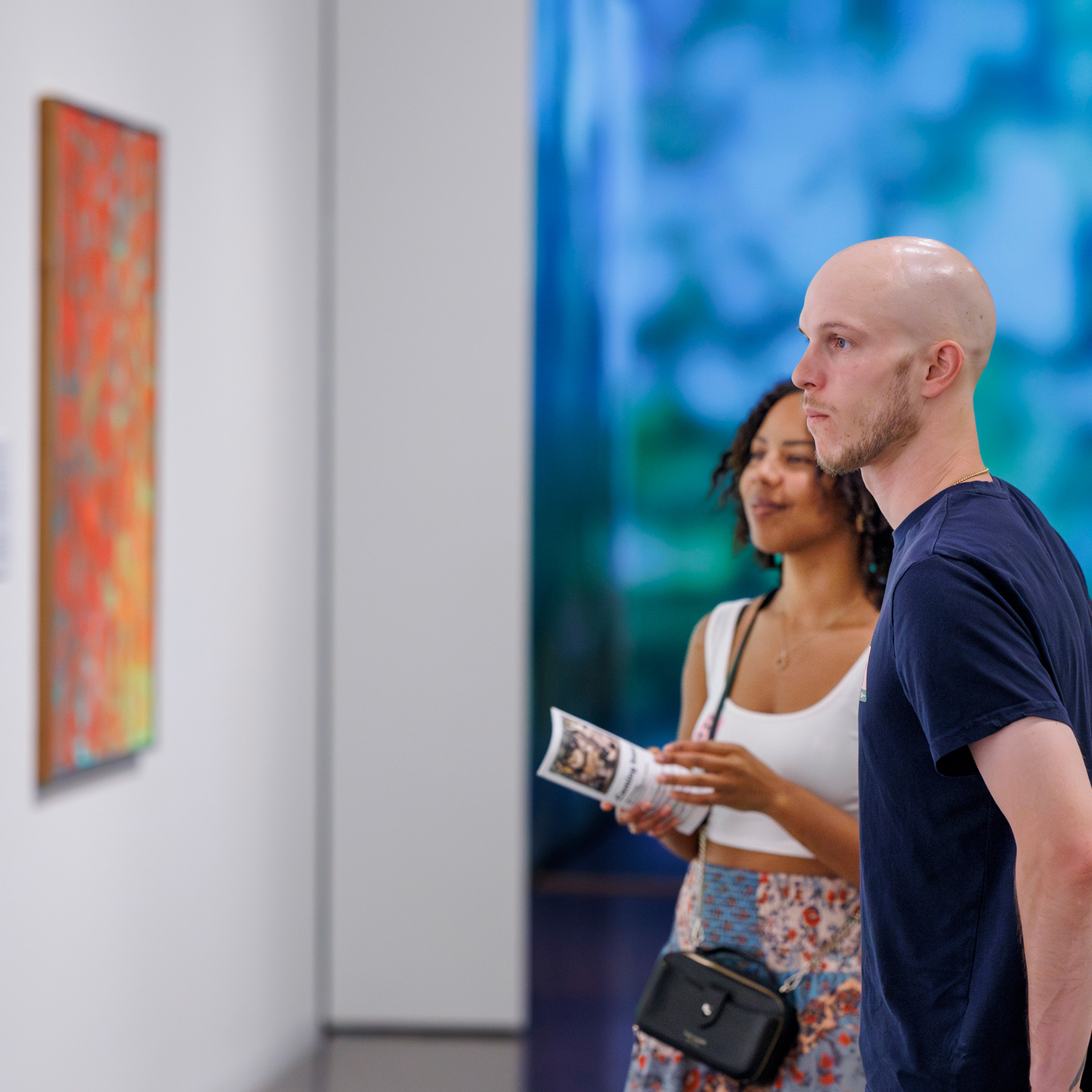 a black woman and a white man look at a colorful abstract painting on a gallery wall