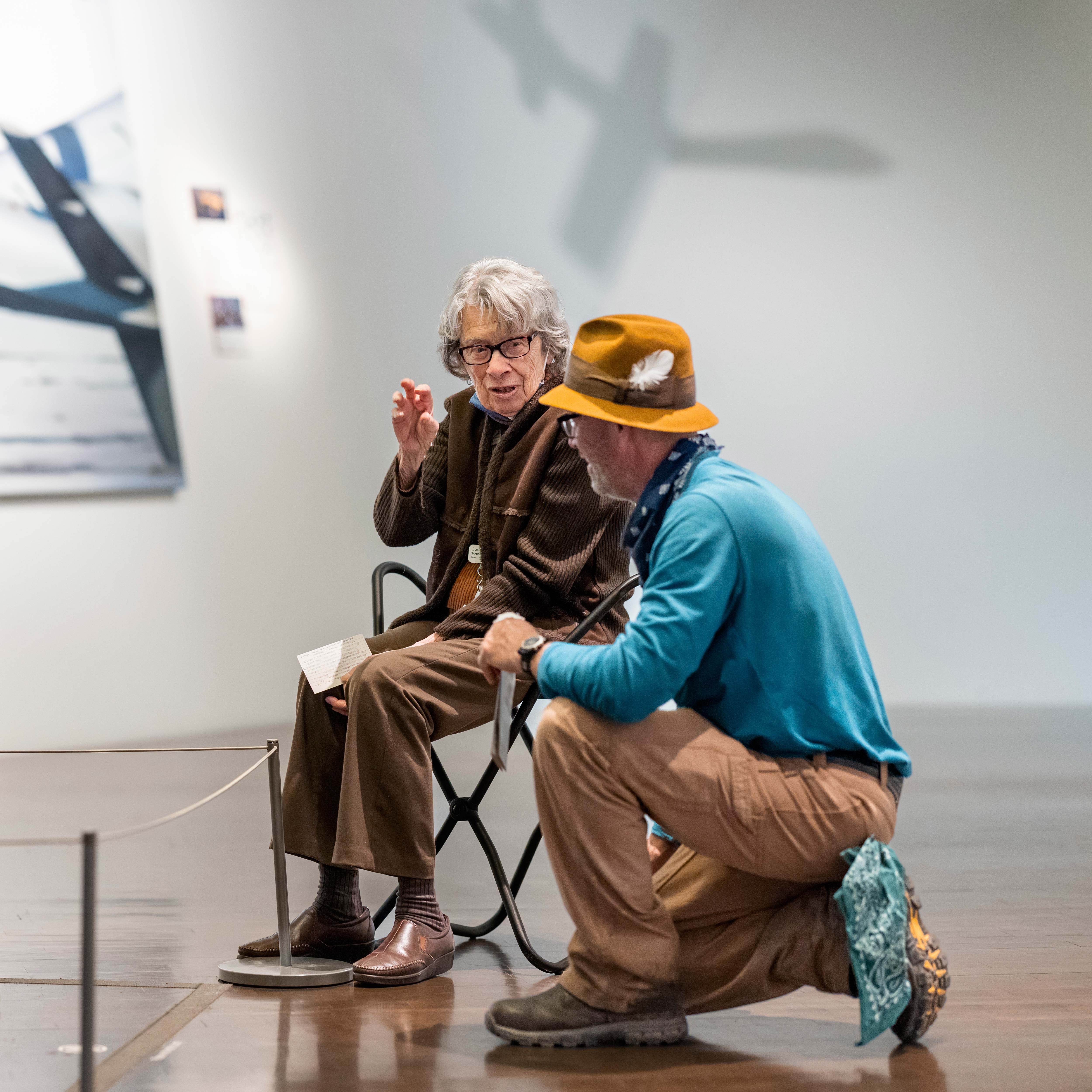 Docent taking a knee to discuss a piece of art with a seated elderly visitor