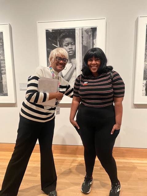 A Black woman with gray hair next to a young Black woman stand side by side in front of black and white photos in the exhibition.