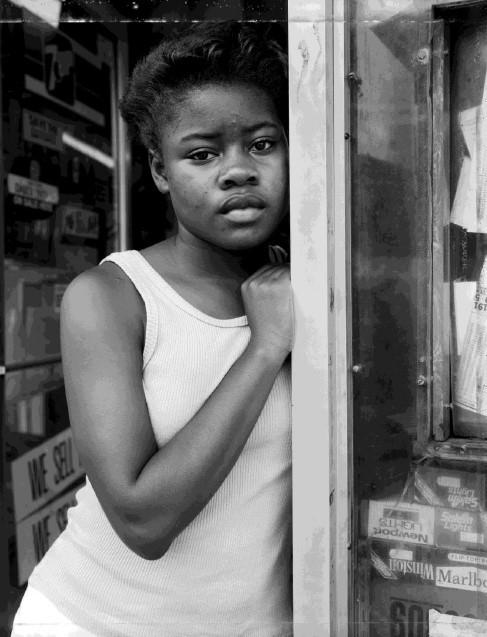 A black and white photograph of a young African American woman wearing a white tank top. She leans against the doorway of a store, looking out at the viewer.