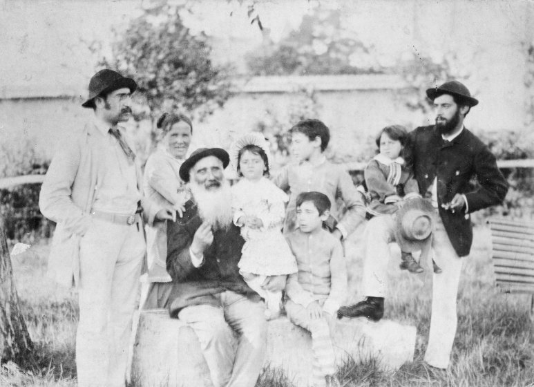 A black and white photo of a family outdoors. An older bearded man sits in the center holding a child, surrounded by adults and children standing or sitting. Trees and a building are in the background.