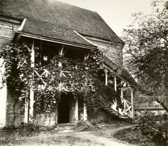 A black and white image of a house covered in ivy. There is a staircase leading across the house to a covered landing on the second floor. There is an entryway underneath the covered second floor entrance.