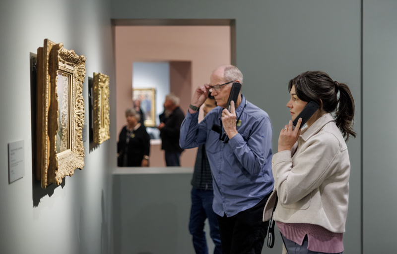 Man and woman holding up audio guide wands to their ears
