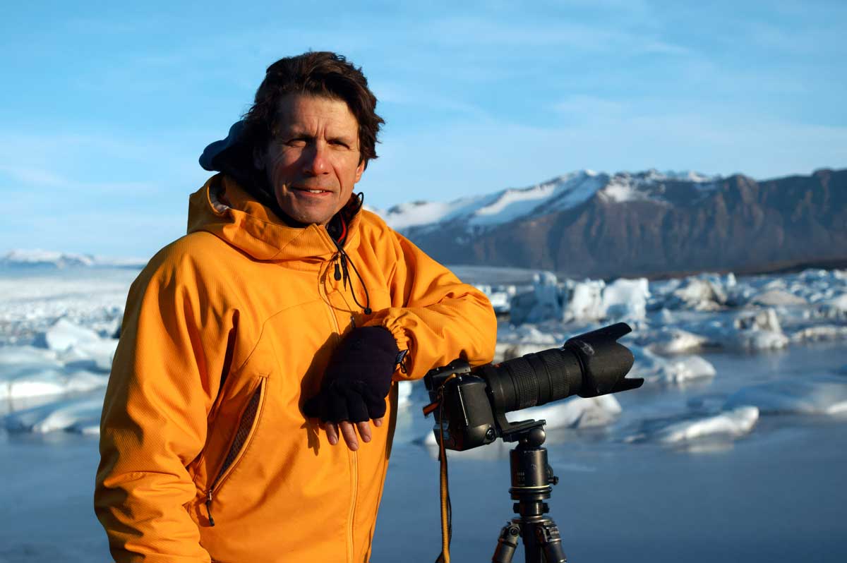 James Balog leans against a camera tripod in an arctic environment. Small icebergs float on calm water in the background.