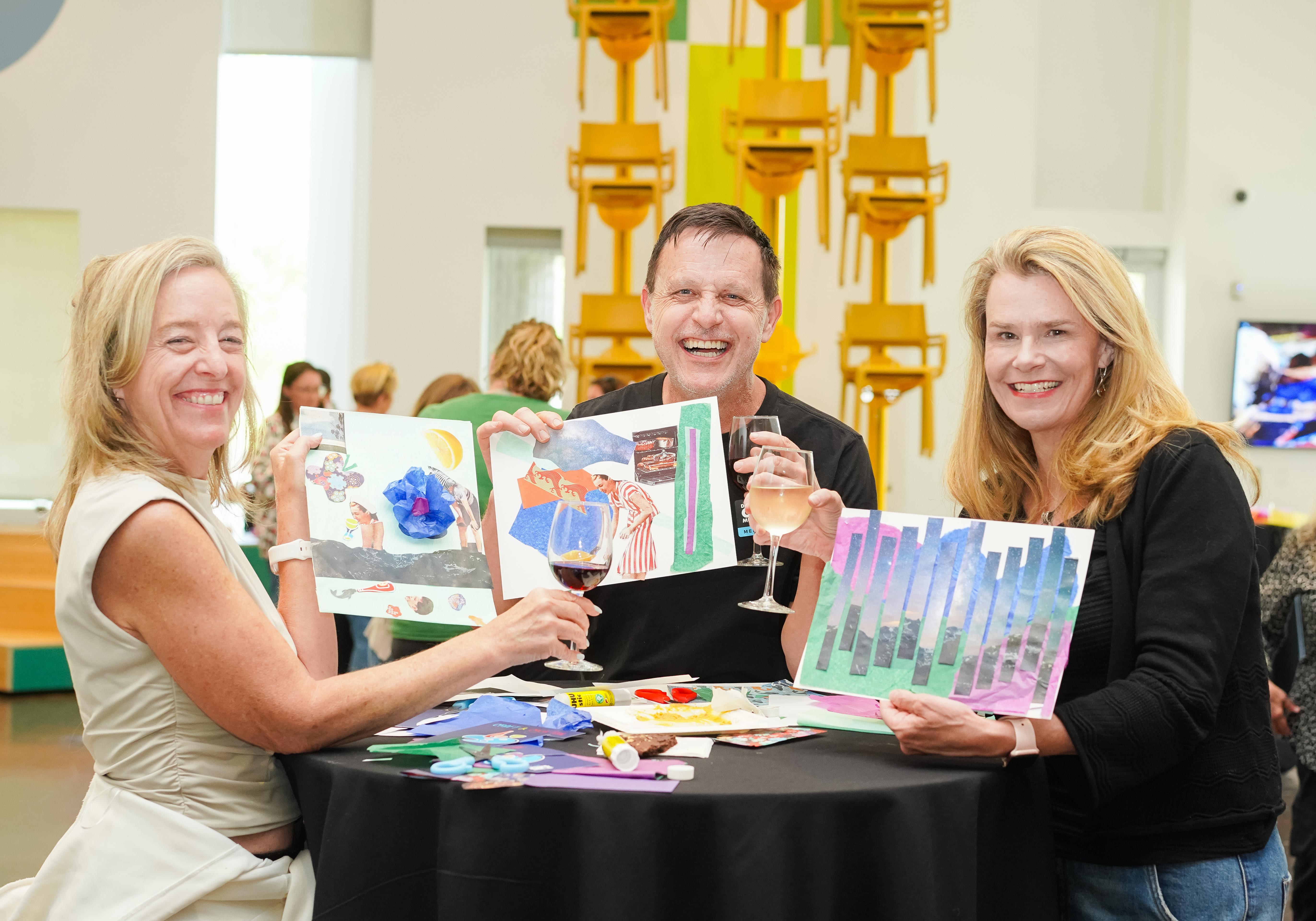 Two women and a man holding up their artworks and clinking their wine glasses
