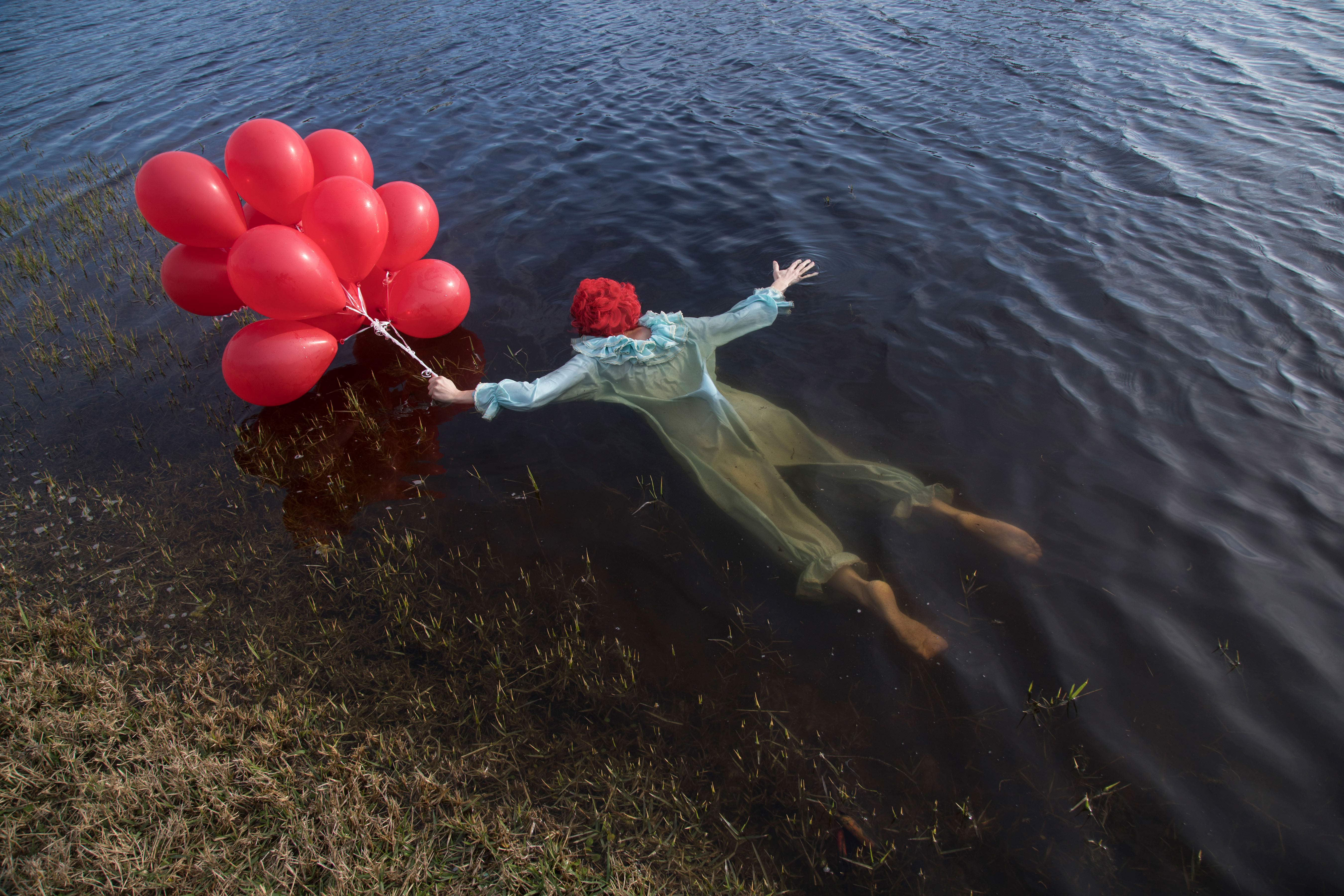Person dressed as a clown lying face down in water, holding a pack of red balloons