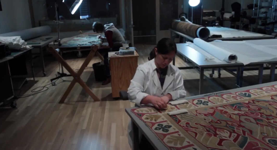 Video still taken from a series of videos documenting the conservation of a Spanish Colonial table cover at the Denver Art Museum. This image shows a textile conservator repairing the tapestry.