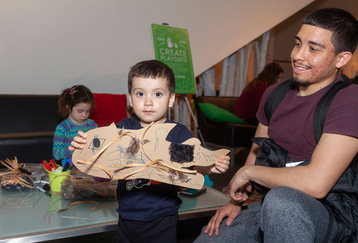 A boy holds up art he made while a man looks on at Create Playdate